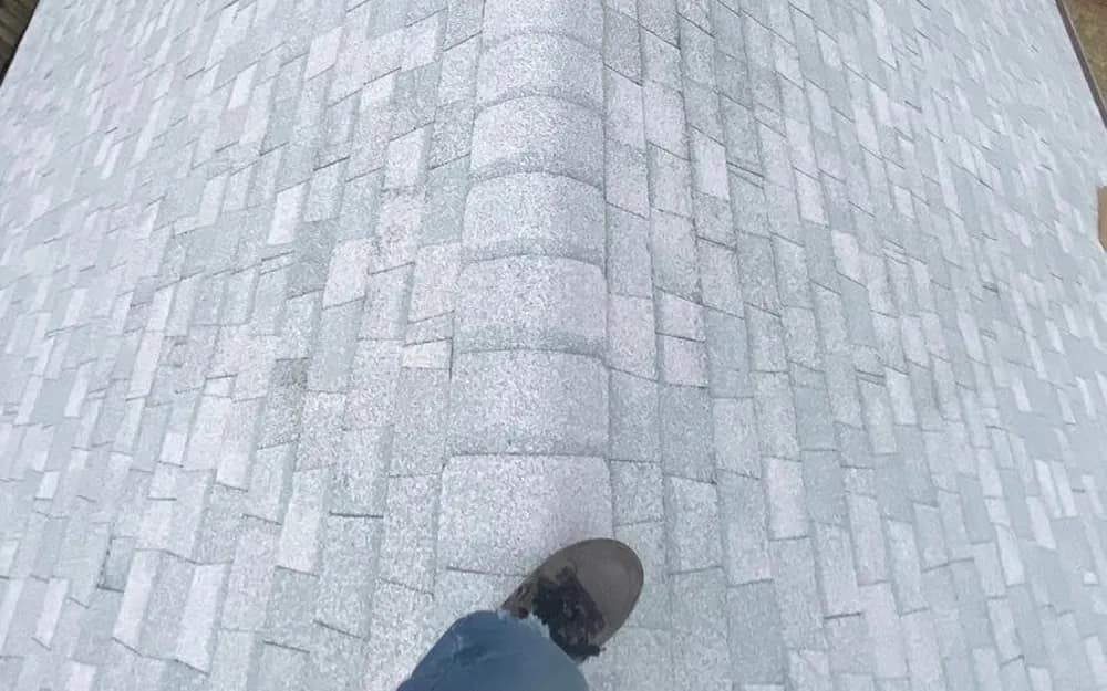 Looking down the ridge of a finished light-gray shingle roof from a roofer's own vantage point, work boot in frame